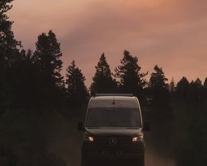 An RV rental camper van travels a dirt road, silhouetted by tall pines against a vibrant sunset sky.