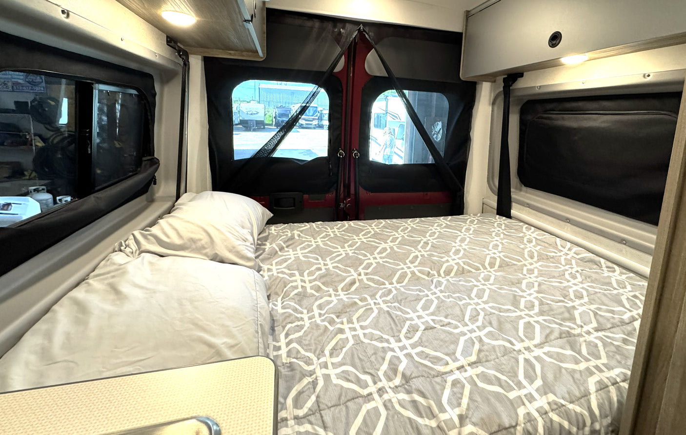 Inside an rv rental, a comfortable bed with grey patterned bedding and windows offering a view of an outdoor vehicle lot.