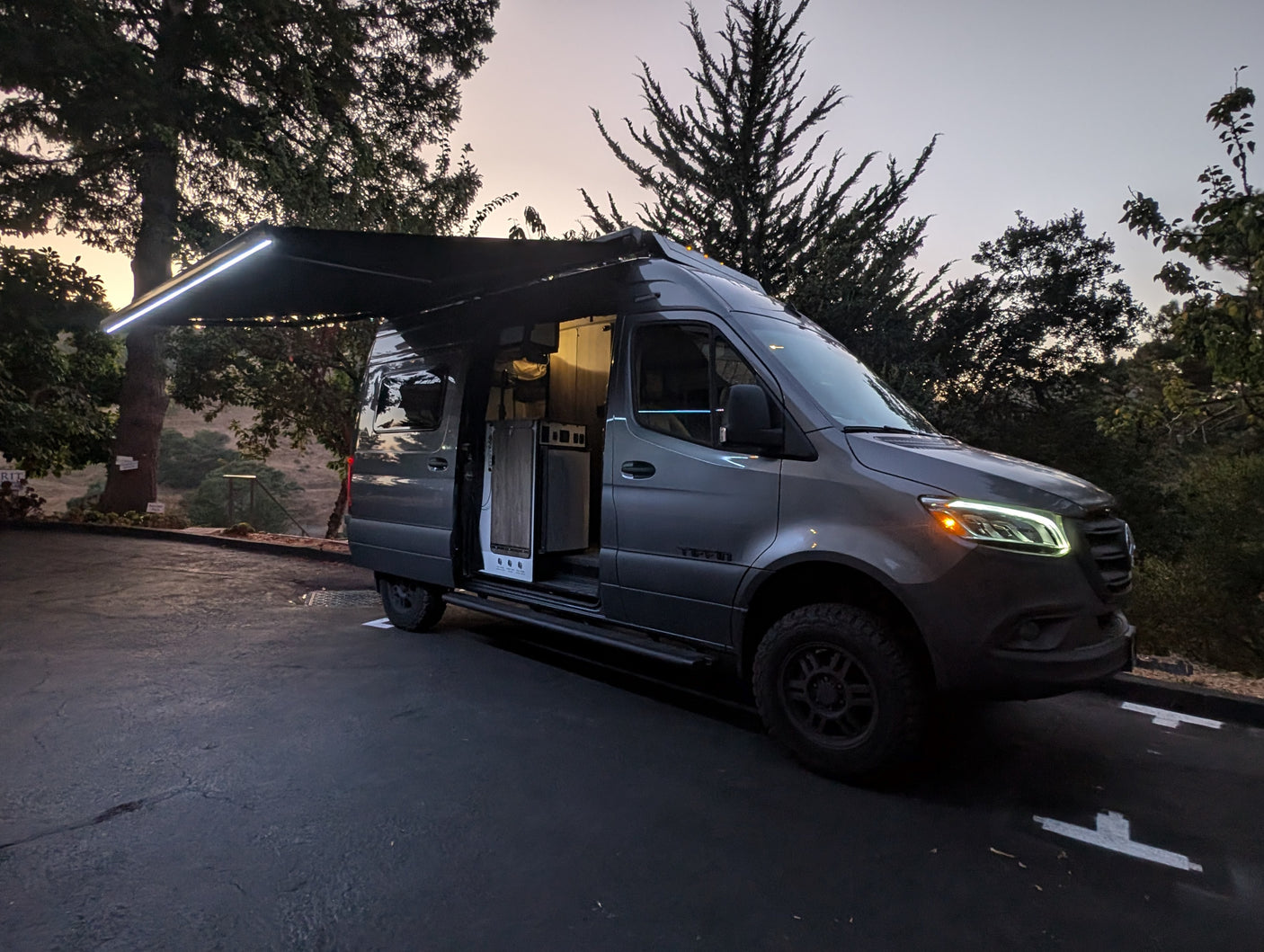 Experience adventure with this gray Mercedes Sprinter rv rental van, featuring an illuminated awning & inviting interior at dusk.
