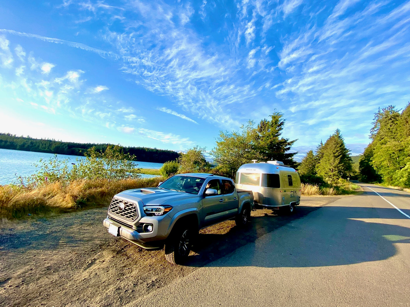 A grey pickup truck towing a silver Airstream rv rental trailer parked by a scenic lake under a bright, cloudy sky.