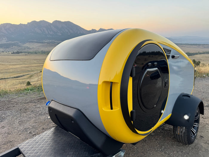Modern yellow & gray teardrop RV rental parked on a gravel path with mountains in the background, ideal for adventure.