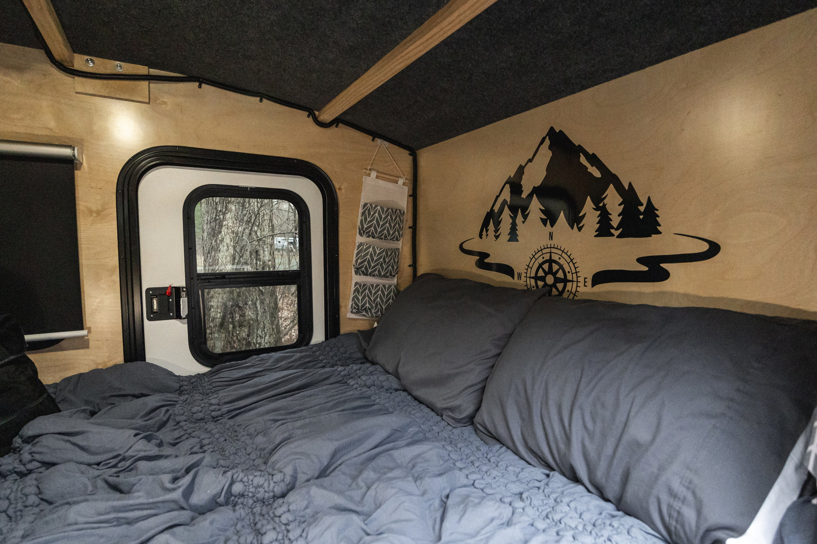Cozy RV rental interior: comfy bed, window with tree view, and scenic mountain decal, ideal for camping and travel.