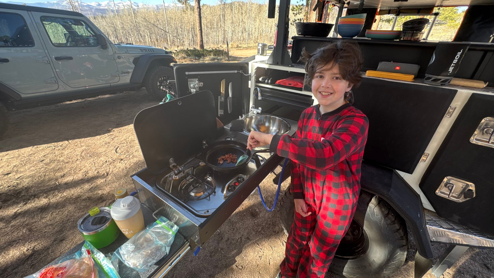 Happy child cooks bacon on an RV rental's outdoor kitchen. Enjoying camping adventure with mountain views.