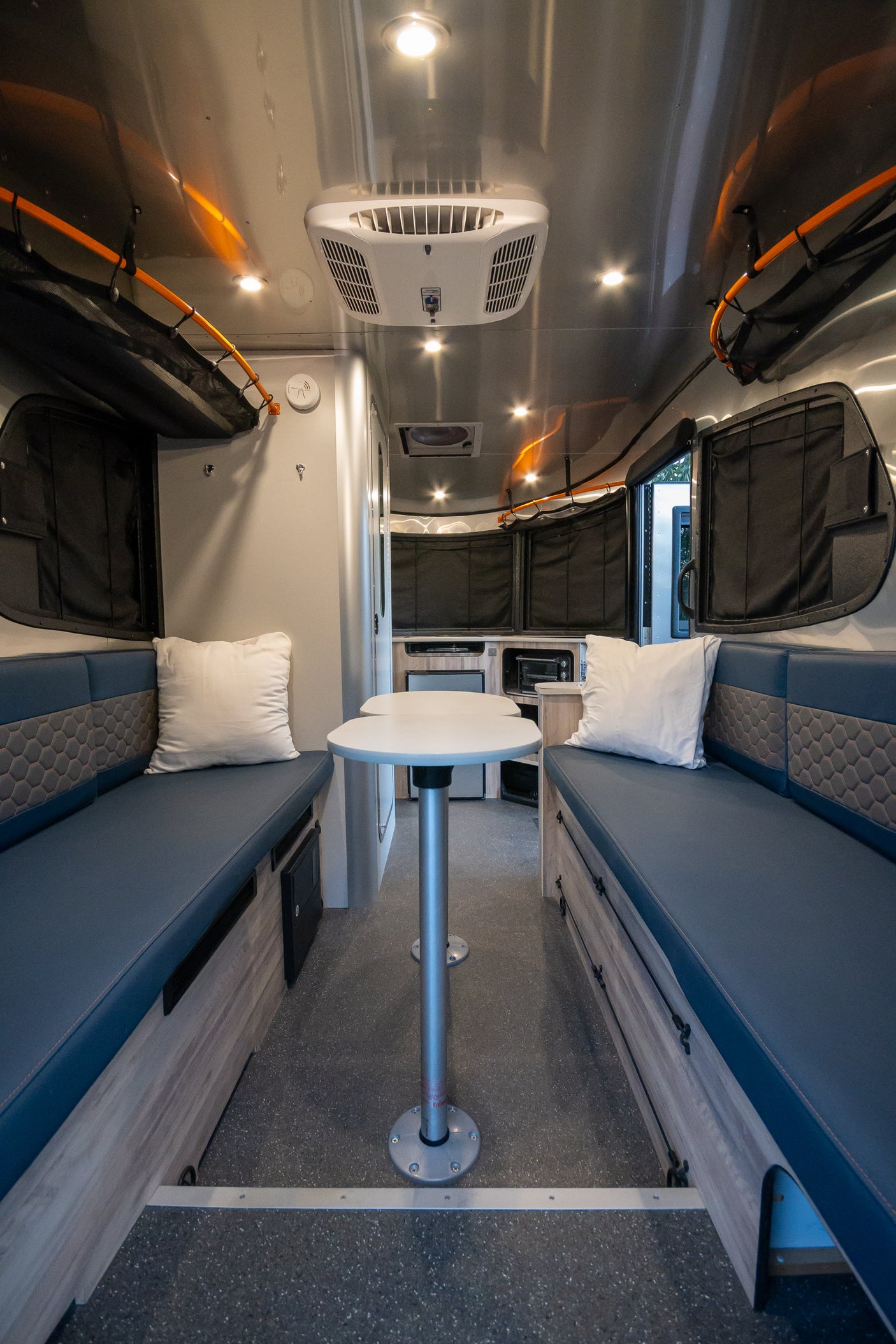 Inside a modern RV rental: a compact dinette with blue benches, central table, and kitchenette. Bright lights illuminate the sleek interior.