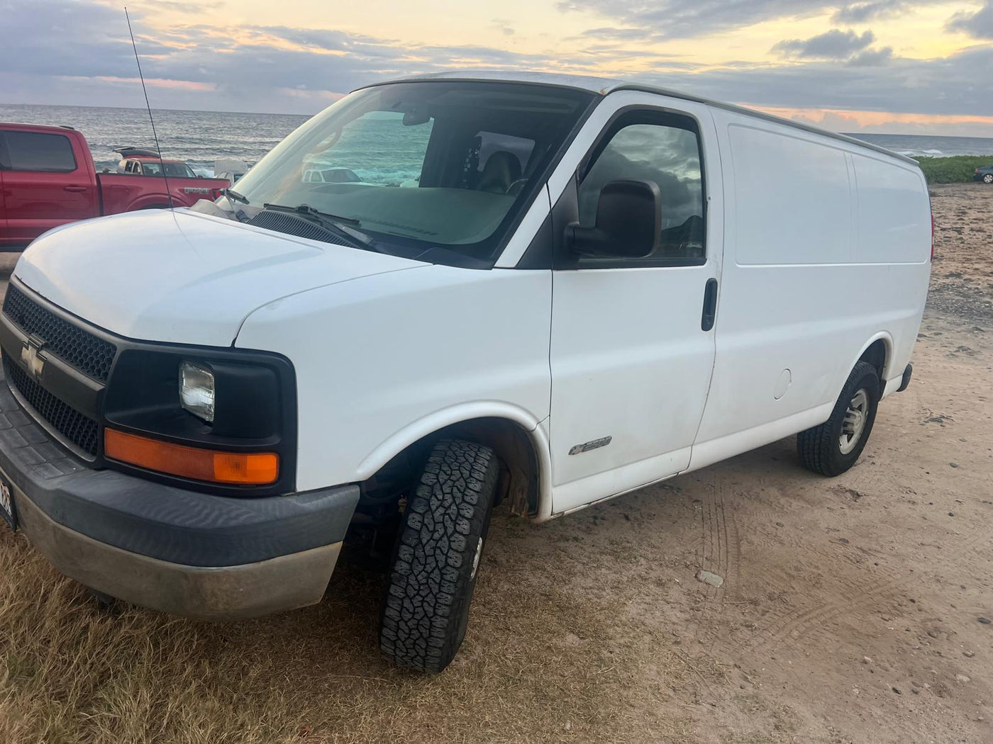 White panel van with all-terrain tires on a sandy beach by the ocean. Ideal for an RV rental or beach camping trip.|RV rental interior: van bathed in red LED string lights on ceiling & walls — photo 1