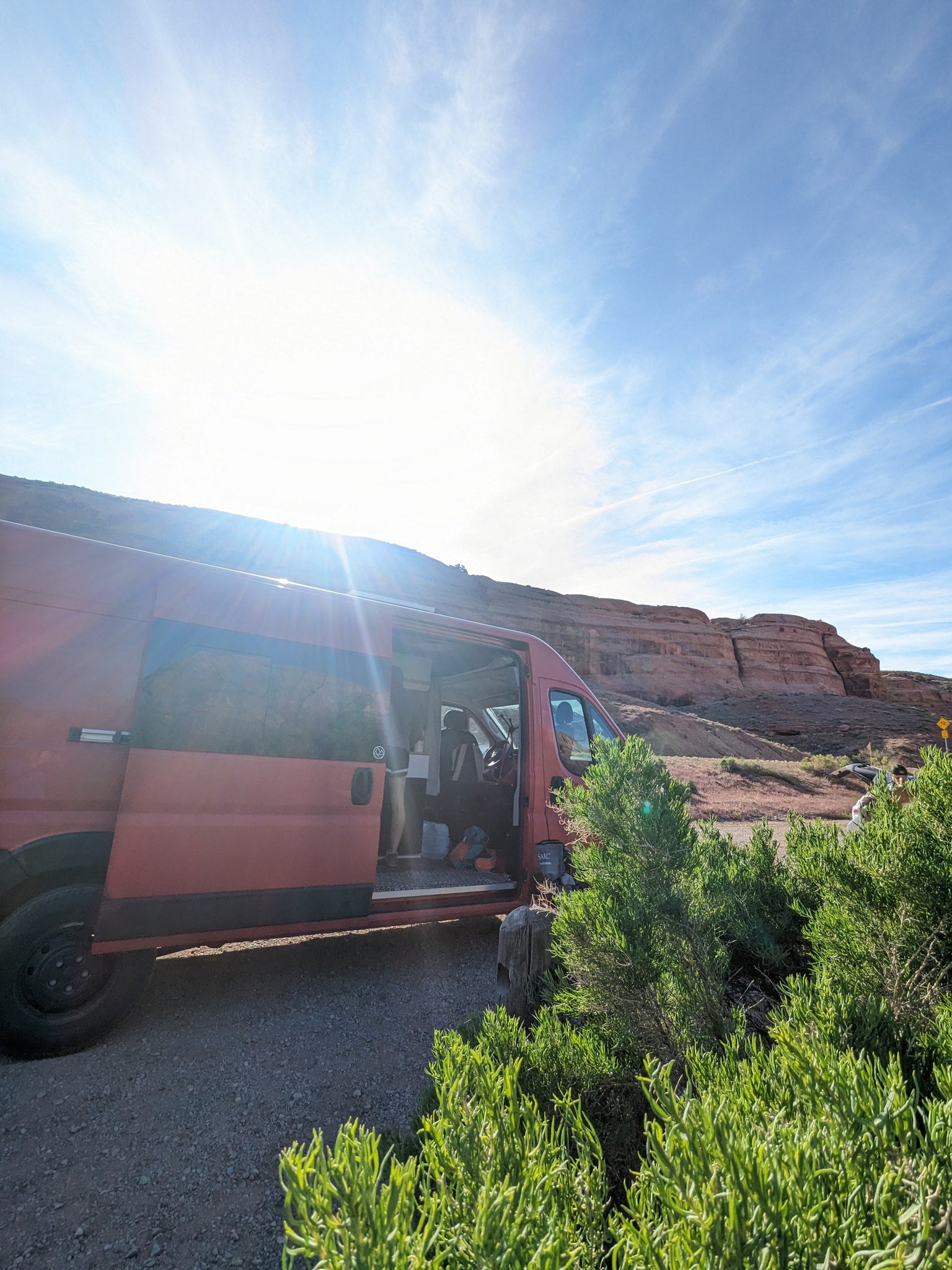 Red camper van parked with open door by red rocky cliffs on a sunny day. Your adventure starts with an RV rental!|This red Ram ProMaster camper van