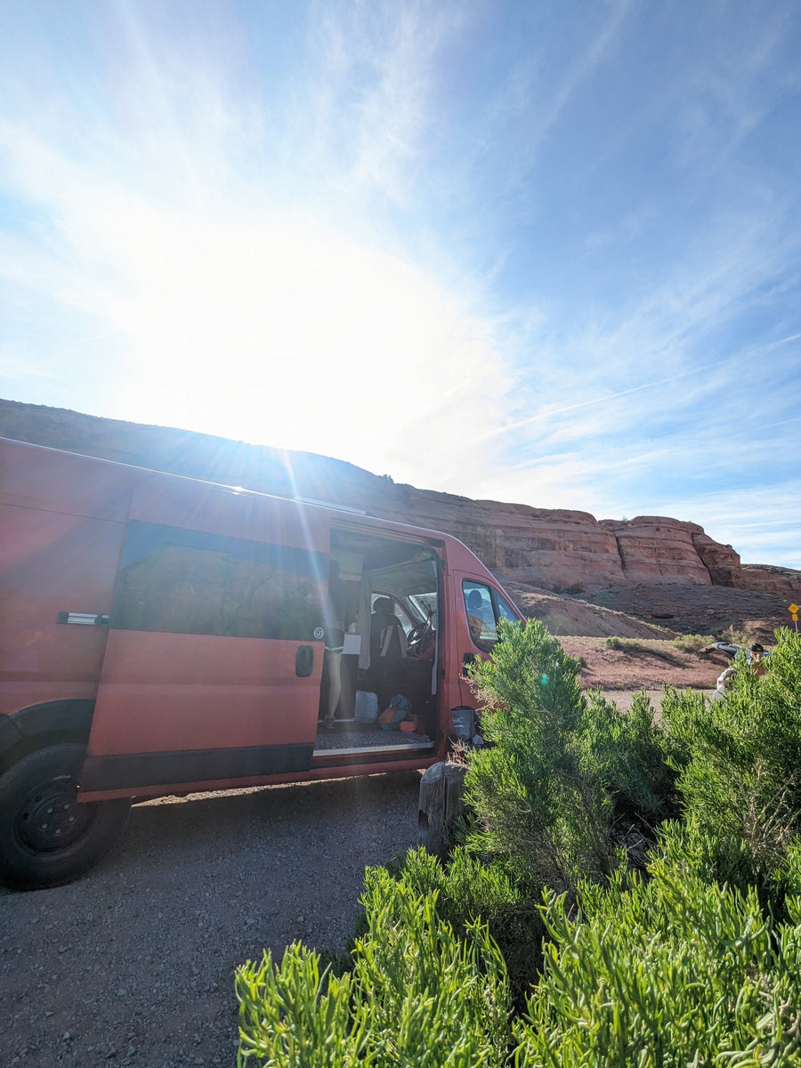 Red camper van parked with open door by red rocky cliffs on a sunny day. Your adventure starts with an RV rental!|This red Ram ProMaster camper van
