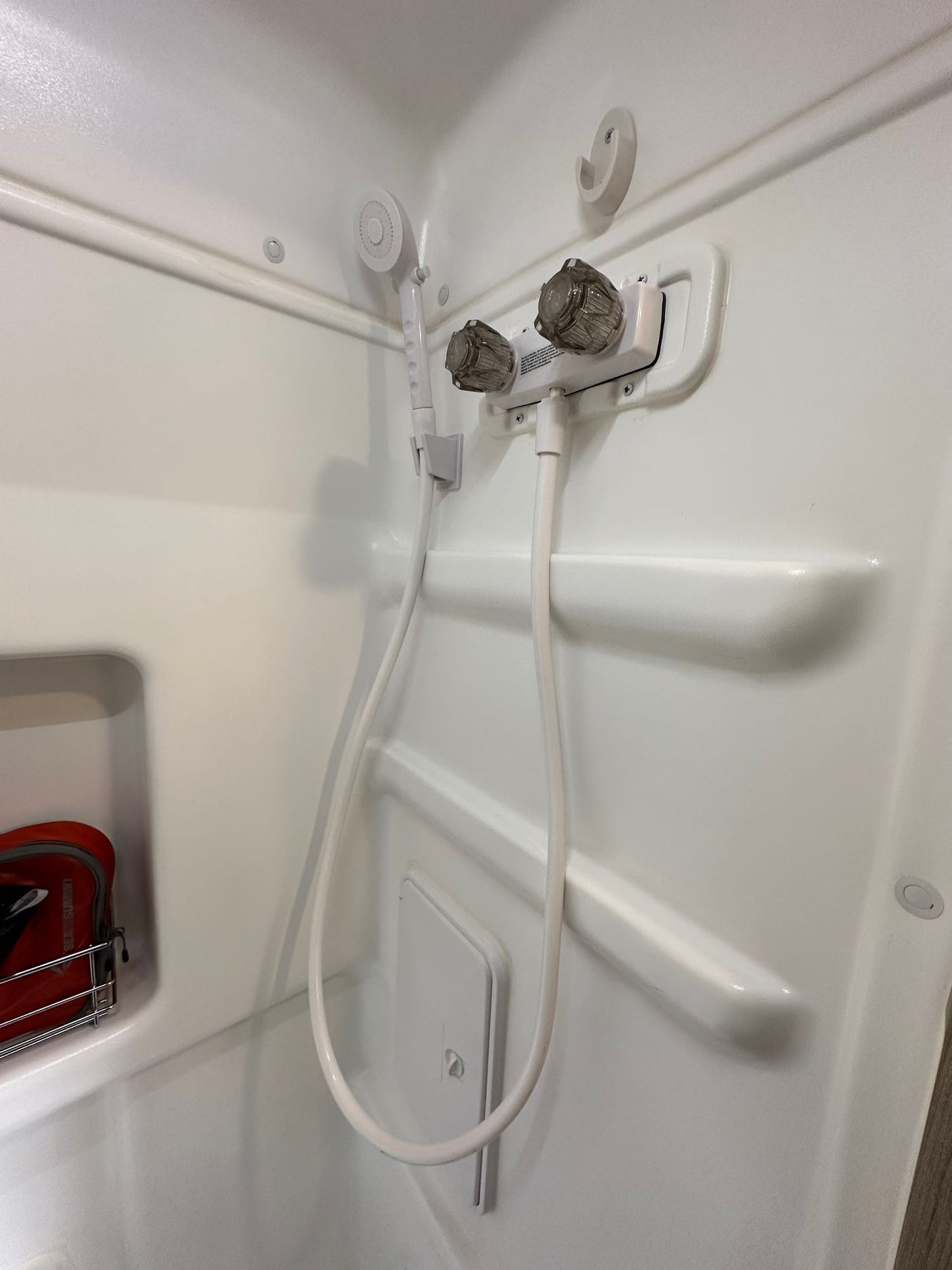 Inside an RV rental: White shower stall with handheld sprayer, dual faucets, wall hooks, and built-in shelving.