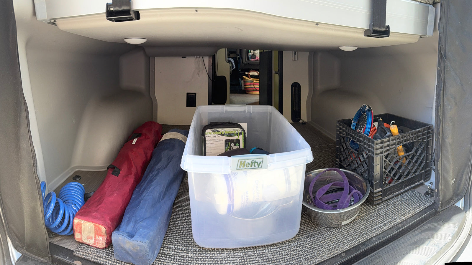 Inside an **rv rental's** rear **storage**, featuring organized **camping gear**: blue hose, bagged chairs, Hefty bin, dog bowl, and crate.