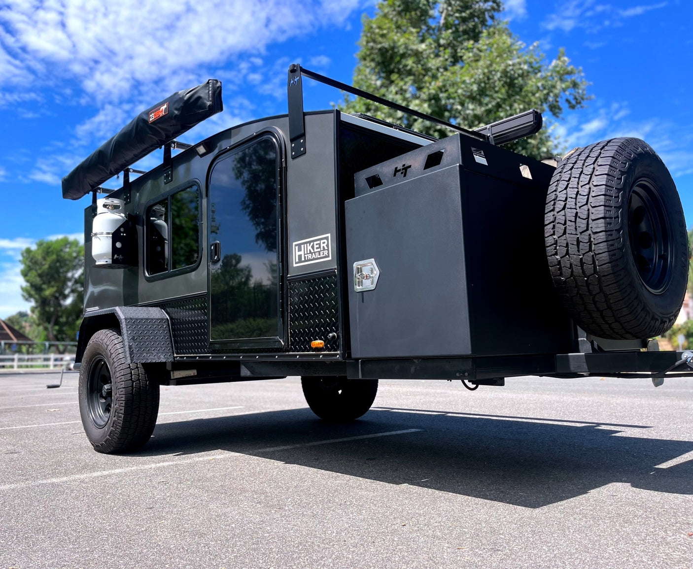 This rugged Hiker Trailer, a dark overland camping trailer with an awning and spare tire, is ready for RV rental adventures.