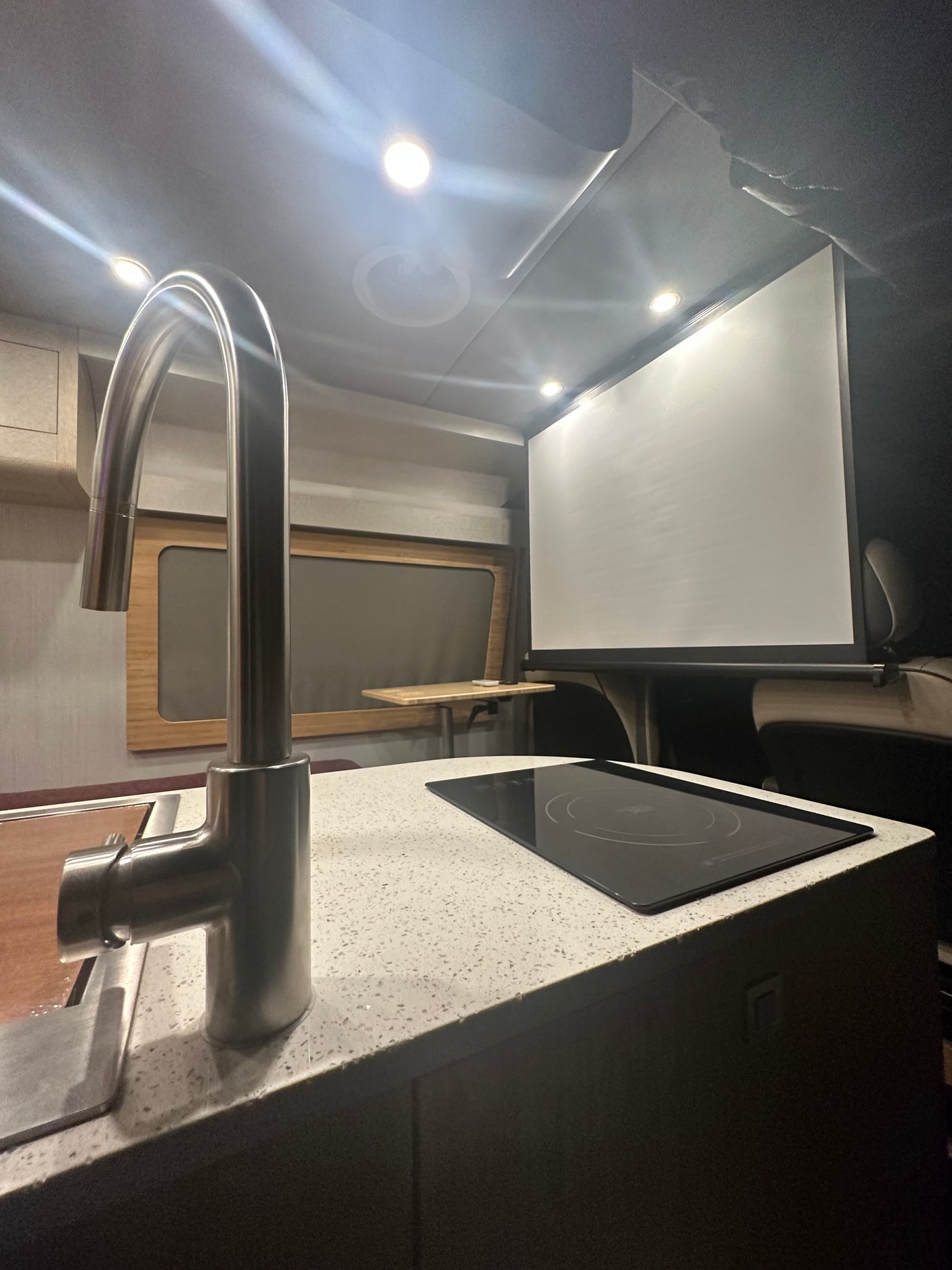 Modern RV rental interior: kitchen with sleek faucet, induction cooktop, and large projection screen for entertainment.