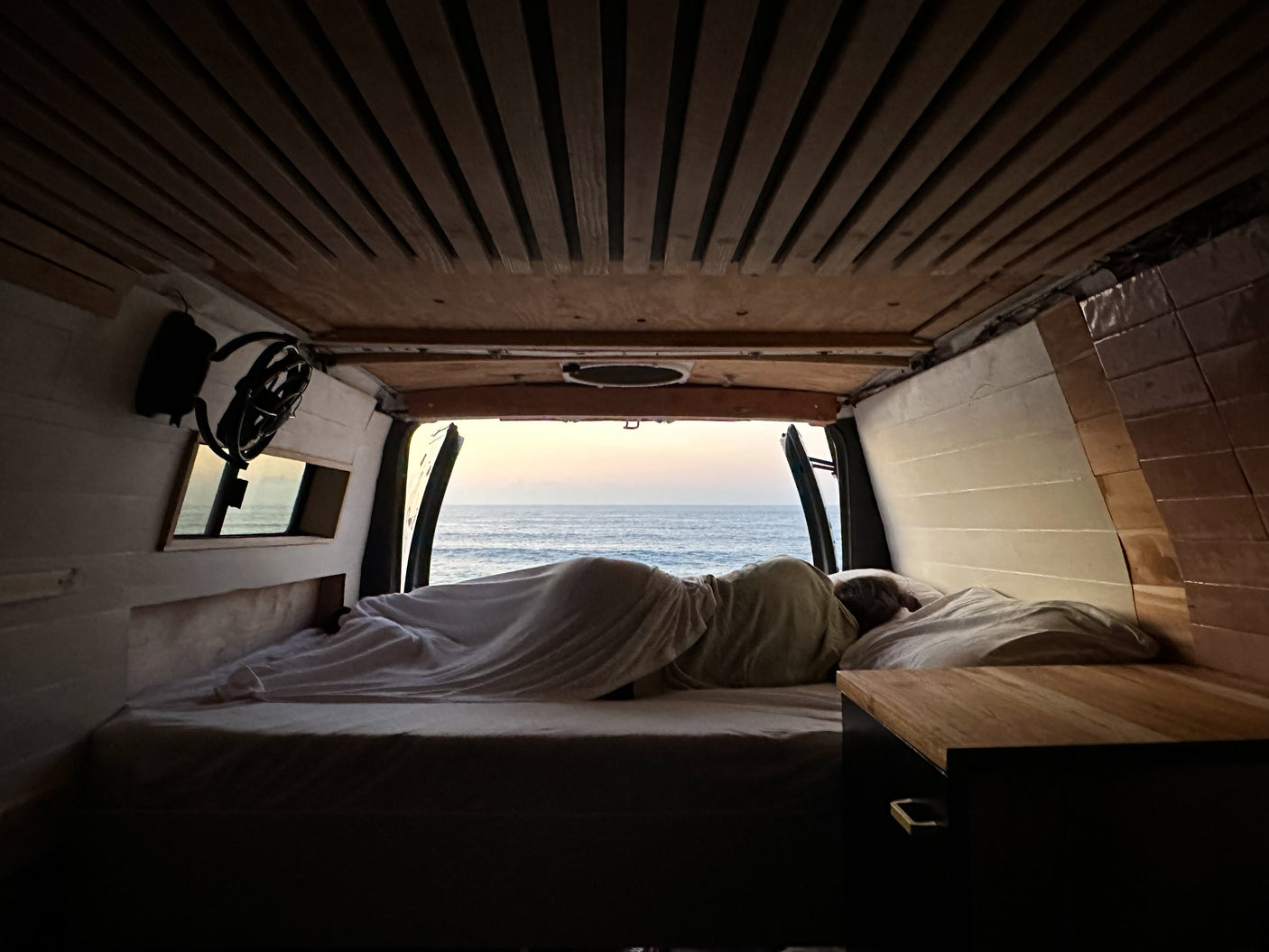 Experience an RV rental: cozy camper bed with a person sleeping, overlooking the ocean at sunrise/sunset.