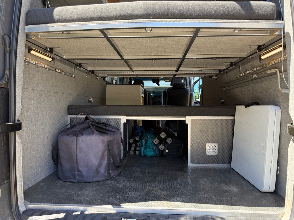 Inside an RV rental, the rear cargo area features an elevated bed, ample storage, side lights, a large black bag, and a white folding table.