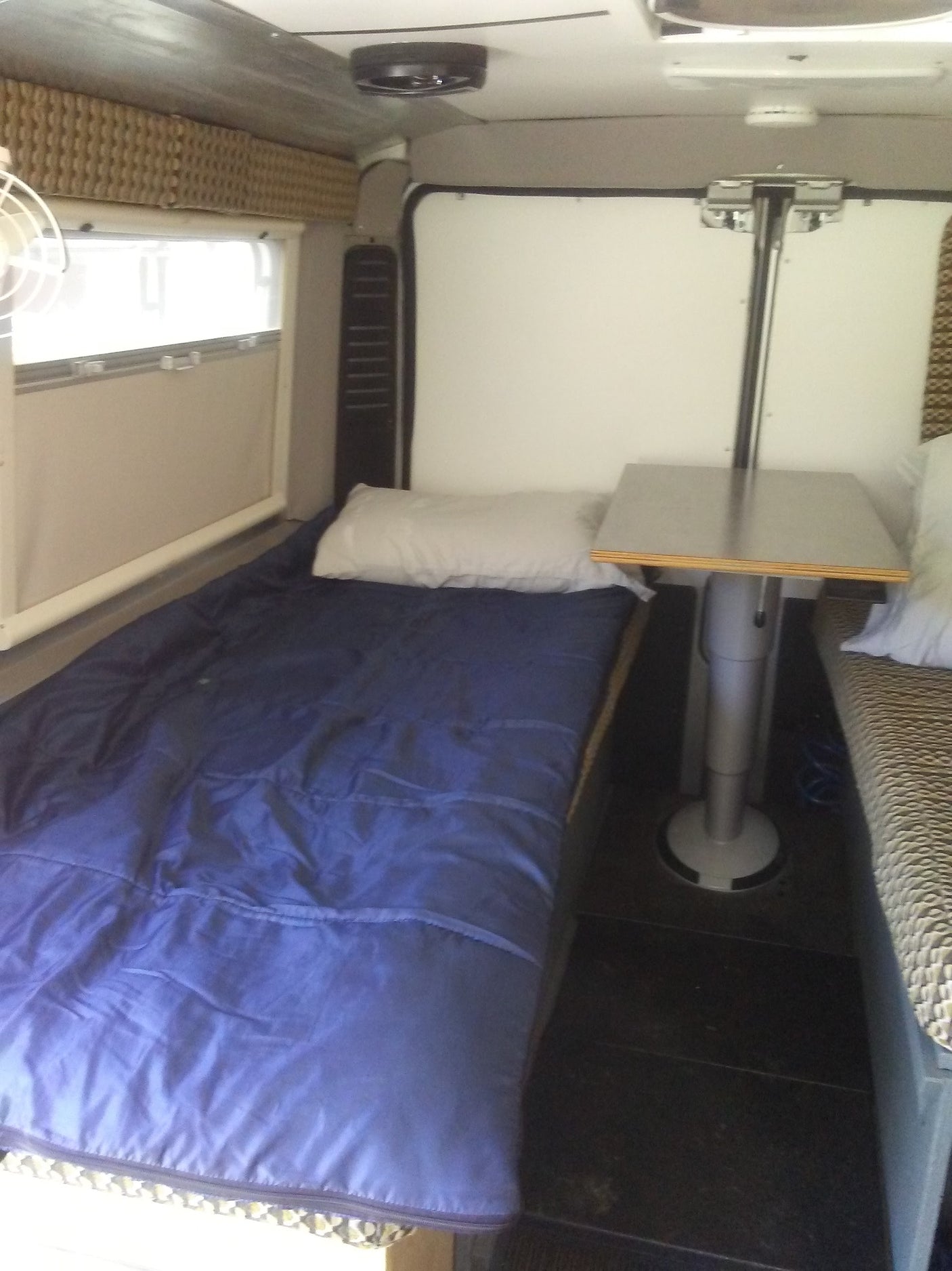 Cozy RV rental interior: A bed with blue bedding, white pillow, and a unique convertible table with a pedestal leg. Window visible.