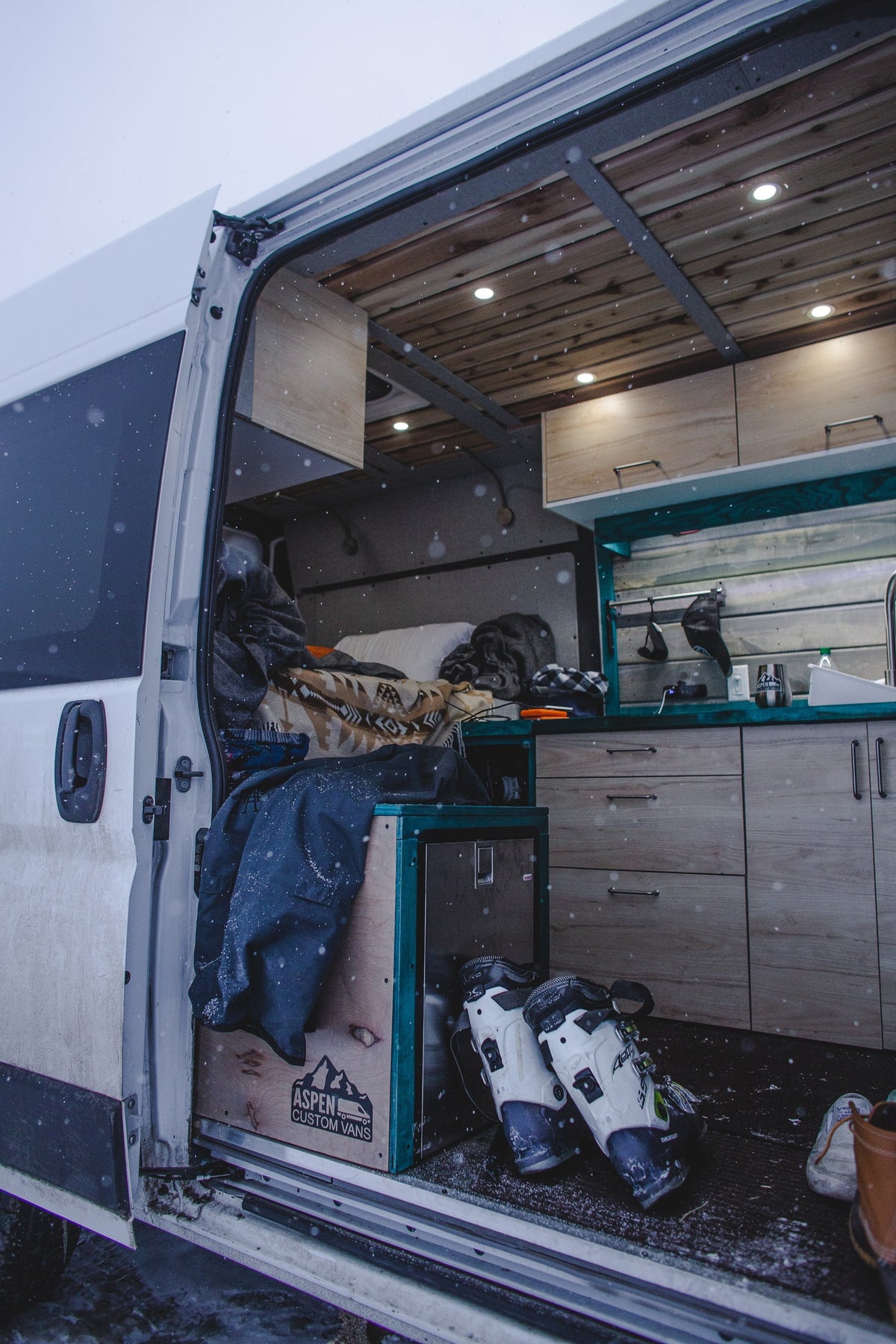 RV rental ready for winter! Cozy custom camper van interior with bed, kitchen, and snow-covered ski boots. — photo 11