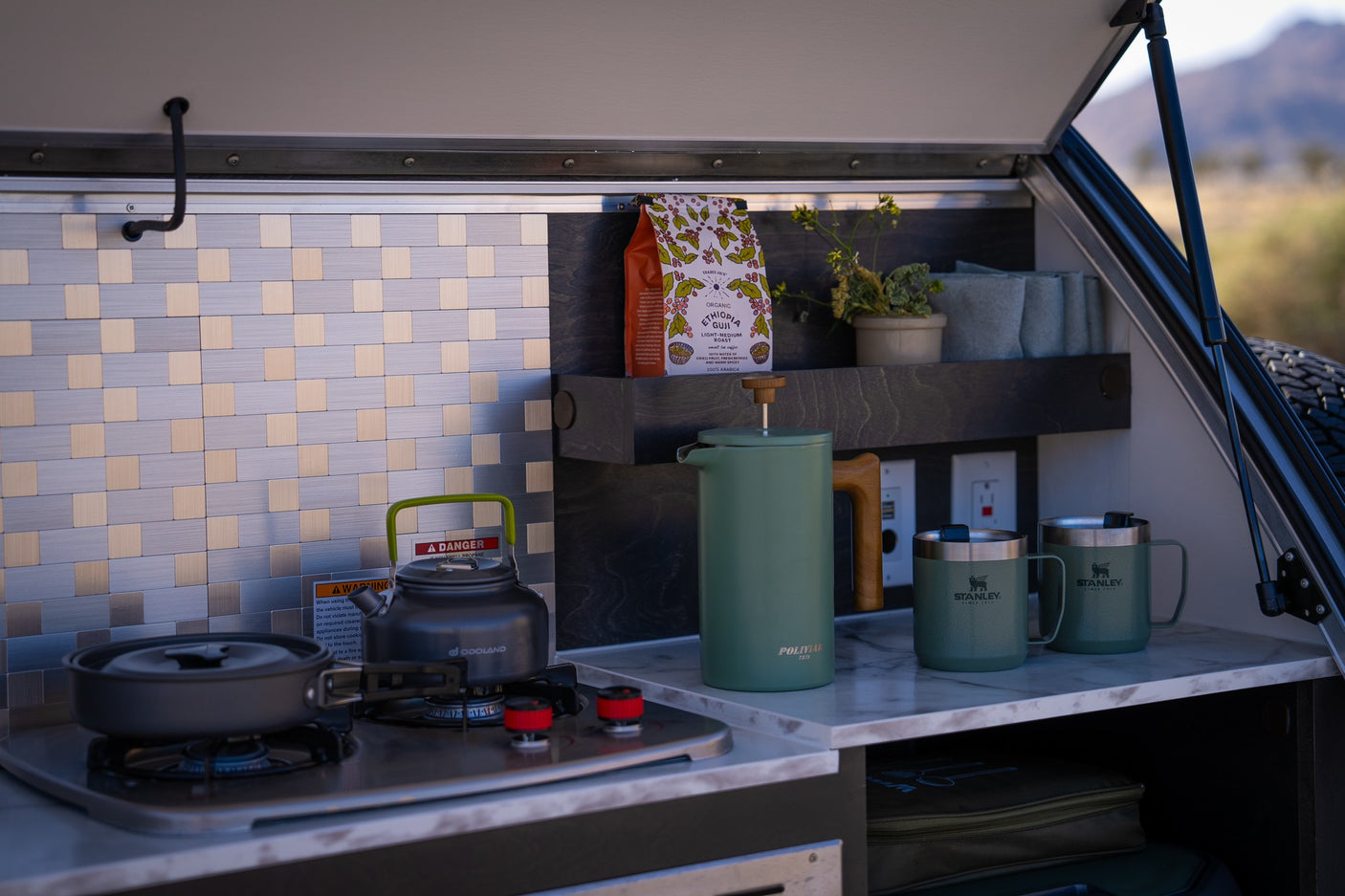 Compact RV rental kitchen with stove, coffee maker, and Stanley mugs, ideal for convenient outdoor cooking.