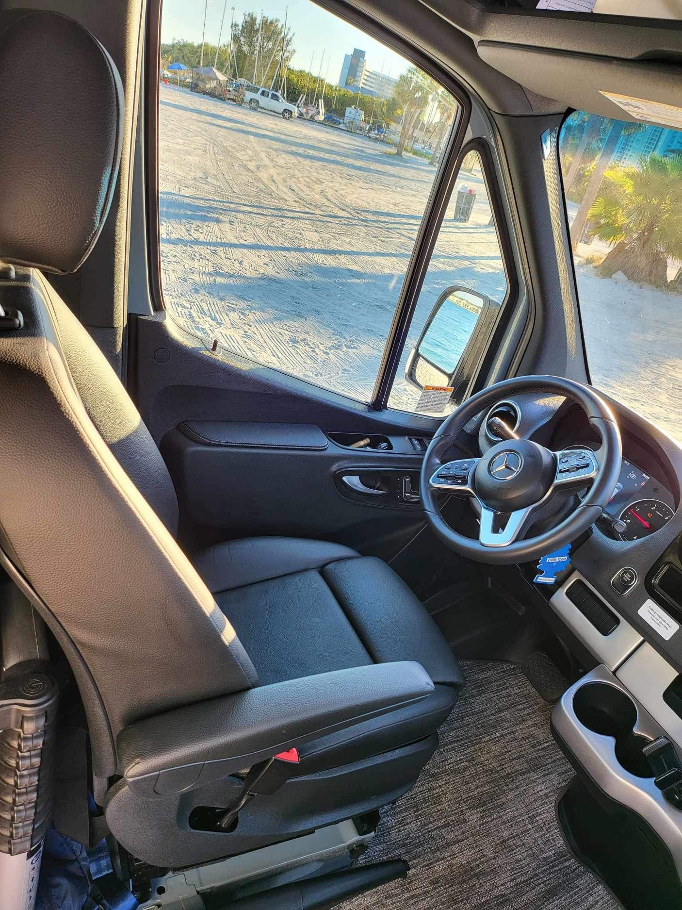 Enjoy your RV rental from the driver's seat! Sunny camper van interior overlooking a sandy beach with parked vehicles and trees.