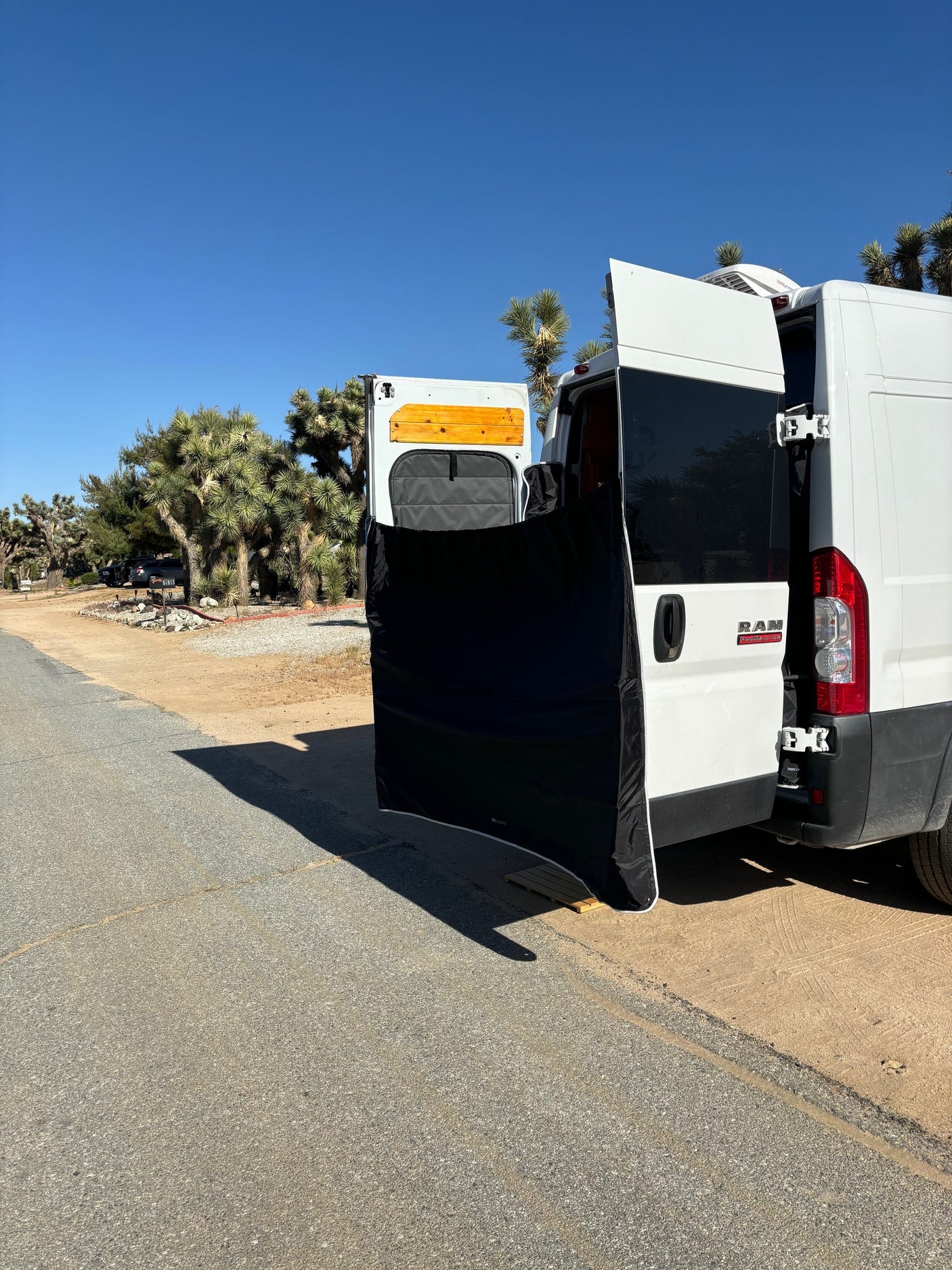 An RV rental van's open rear doors with a privacy screen set up, amidst Joshua trees on a sunny day.