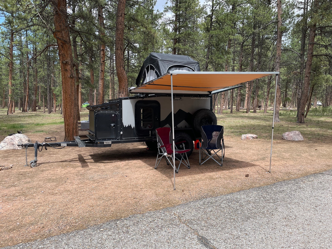 An RV rental overland trailer with a rooftop tent and awning set up in a peaceful pine forest campsite, with two chairs. — photo 9