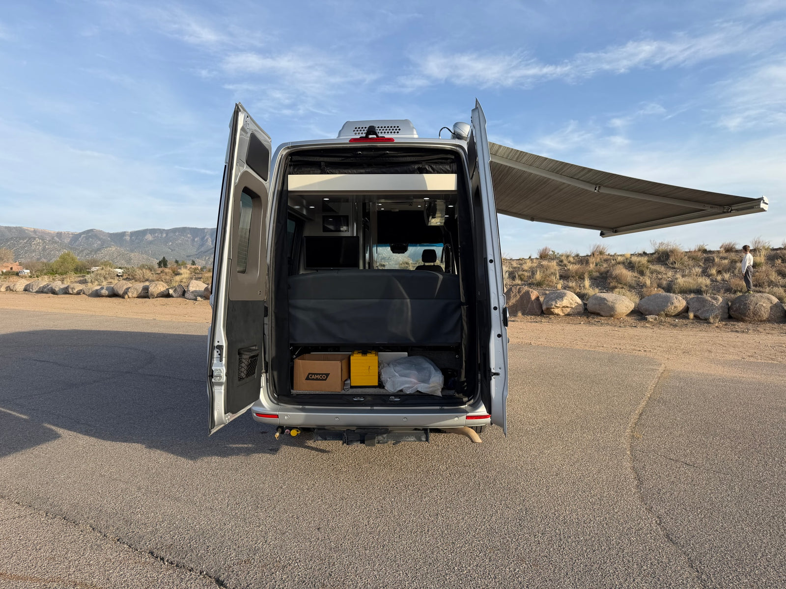 Open back of a silver camper van RV rental, showing interior storage, bed, extended awning, and scenic views.