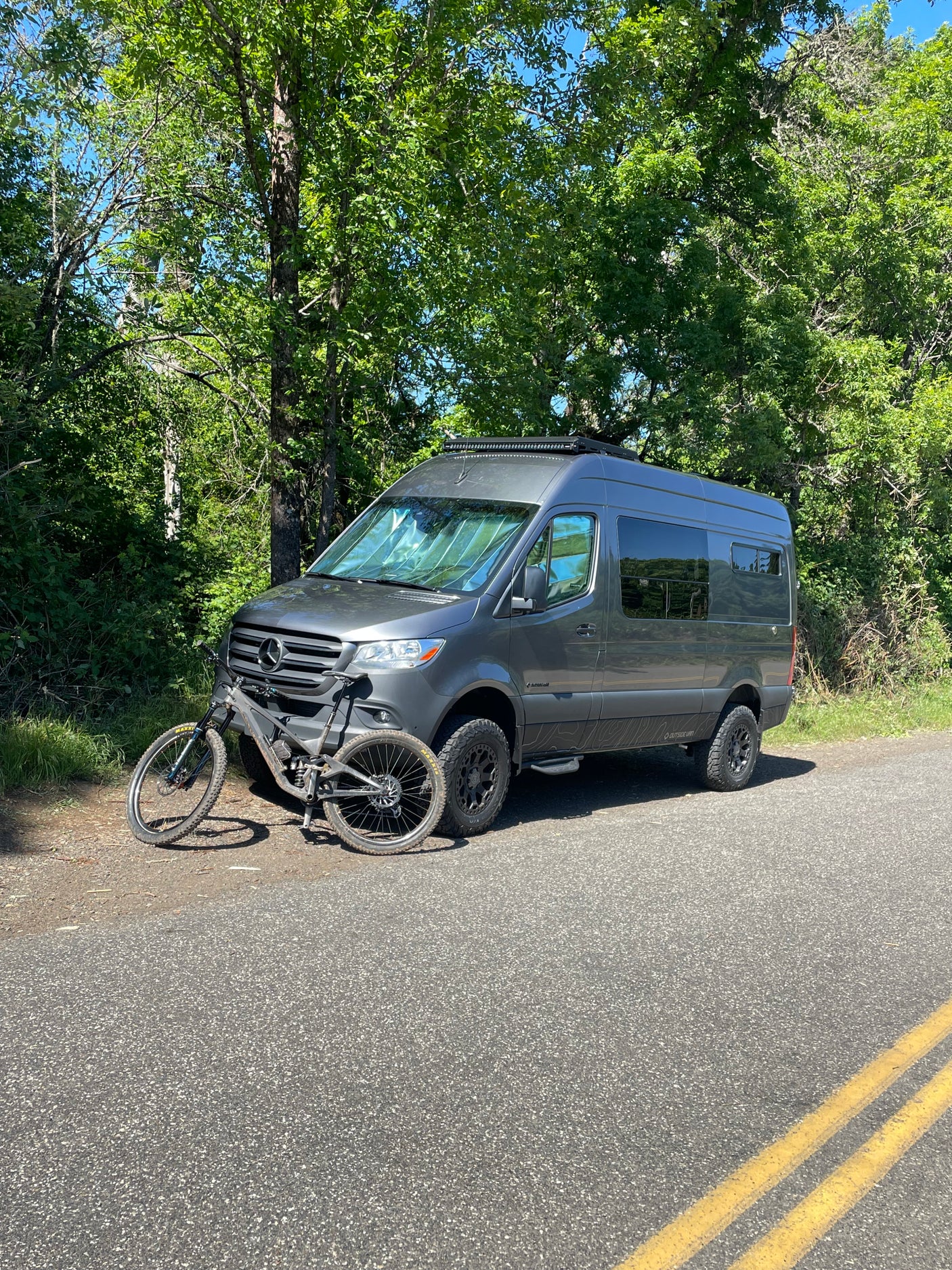 Gray Mercedes camper van (likely an **RV rental**), with a mountain bike, is parked on an asphalt road next to green trees, ready for adventure.
