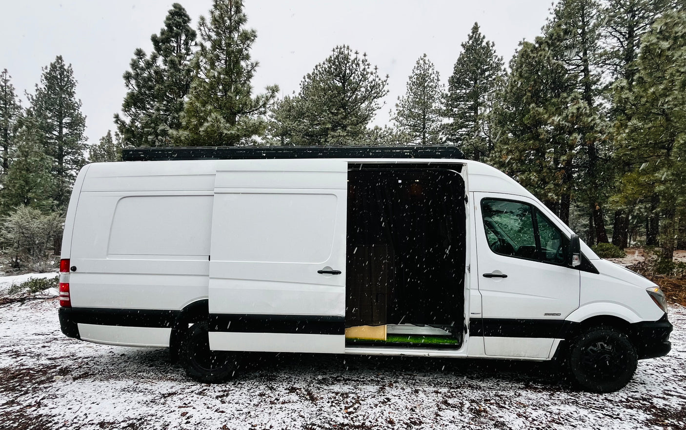 White rv rental van, side door open, parked in a snowy forest with falling snow. Ready for a winter camper van trip.