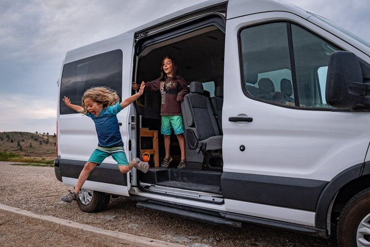 Two excited children leap from a white RV rental van
