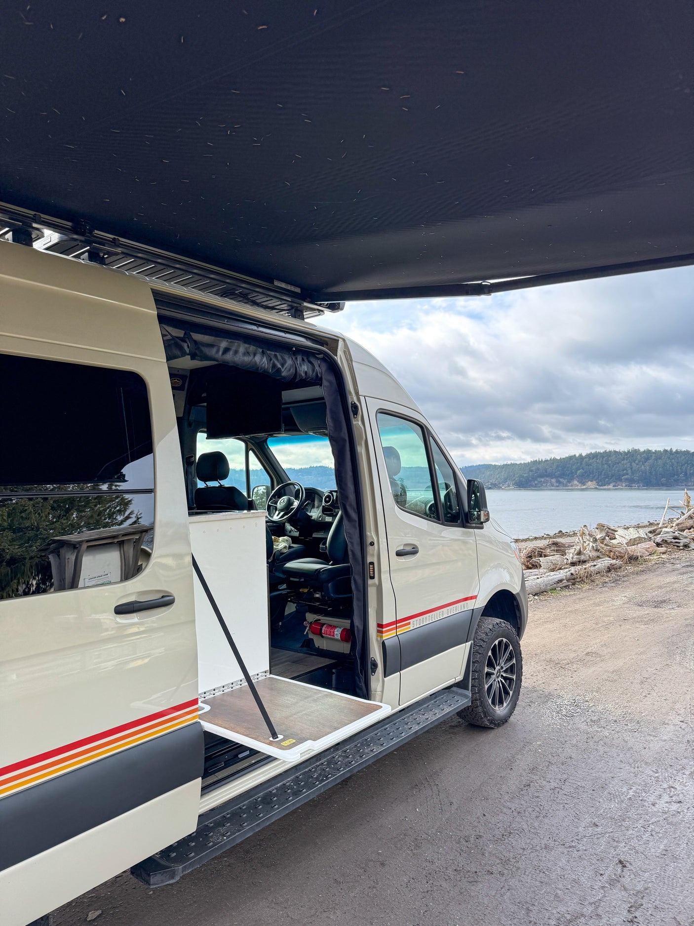 Beige Storyteller Overland camper van, door open with pull-out table, awning deployed by a scenic bay. Your ideal rv rental!