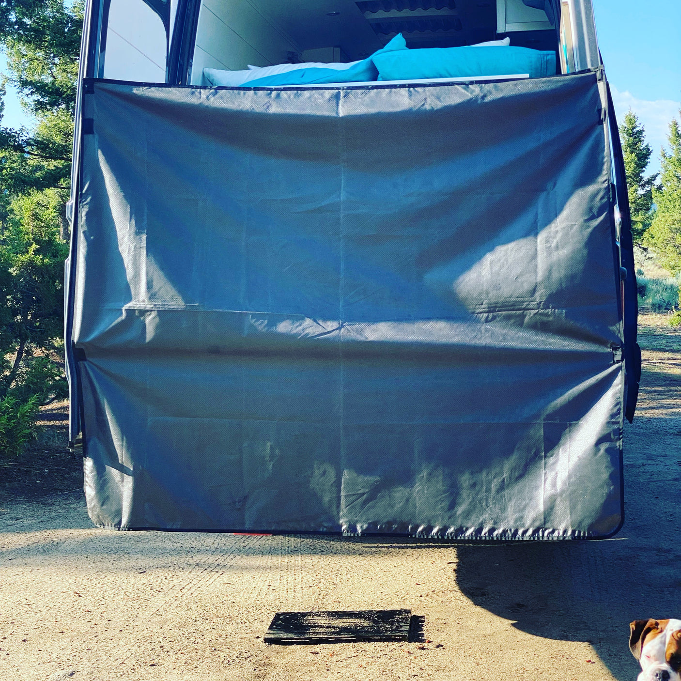 Enjoying an rv rental: rear view of a camper van with a large gray privacy screen, revealing a cozy bed inside. A dog is nearby.