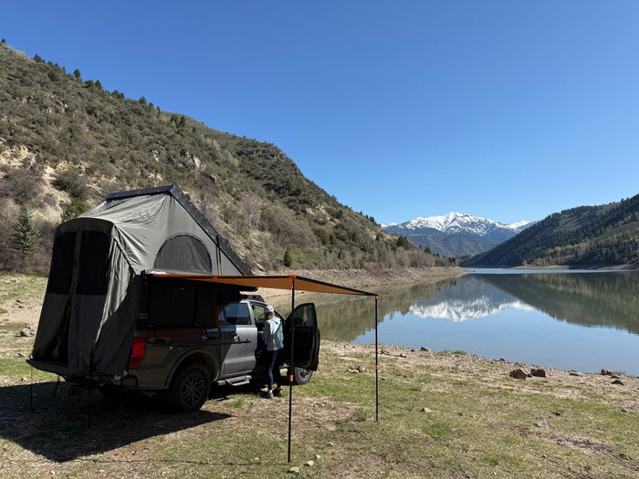 A truck with rooftop tent and awning by a scenic lake, with snow-capped mountains reflected. A perfect outdoor getaway, like an RV rental.
