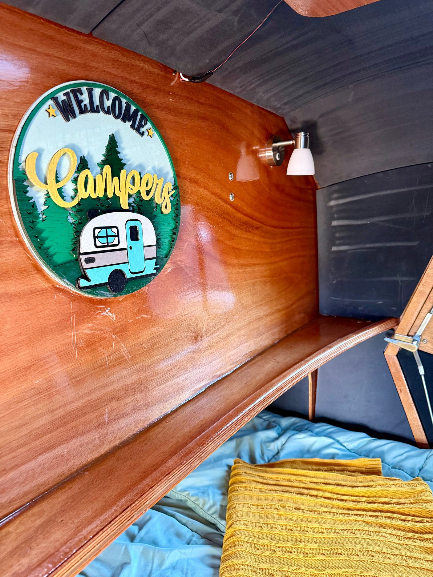 Cozy rv rental interior featuring a "WELCOME Campers" sign on wood paneling, a small light, and a comfy bed.