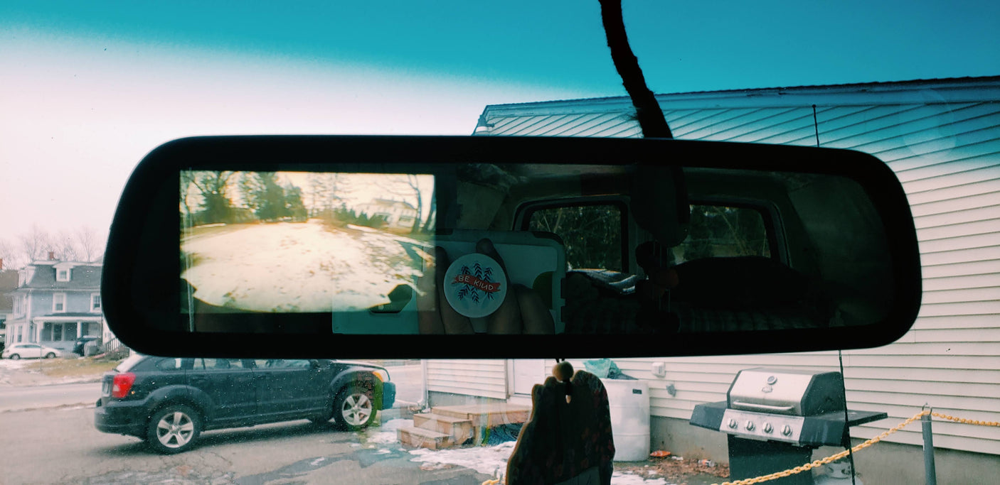 A cozy RV rental view: snowy road in the mirror, "Be Kind" message. Outside, an SUV, house, and grill complete the scene.