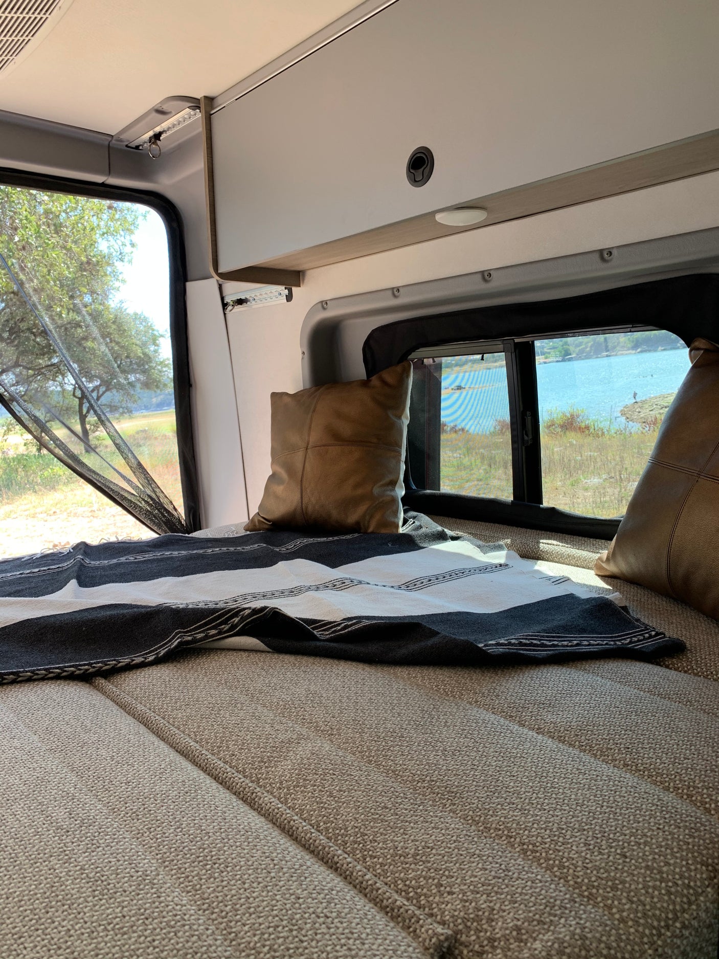 This rv rental offers a comfortable bed with a striped blanket and pillows, overlooking a peaceful lake.