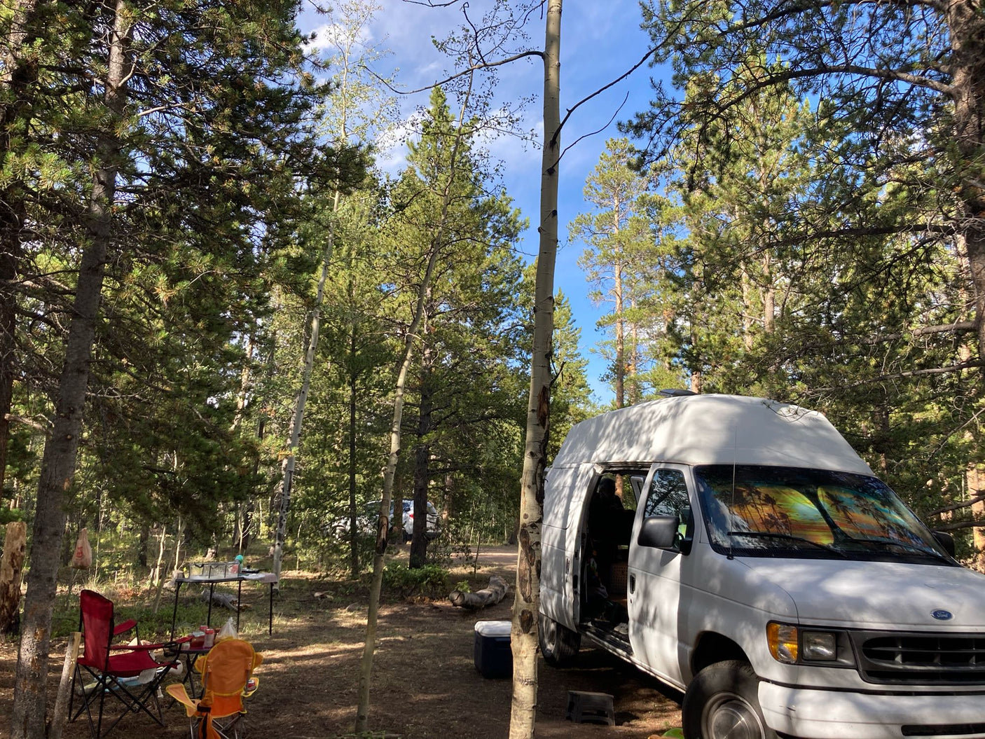 An RV rental van is parked at a serene forest campsite. Chairs and a table are set up, ready for outdoor living.