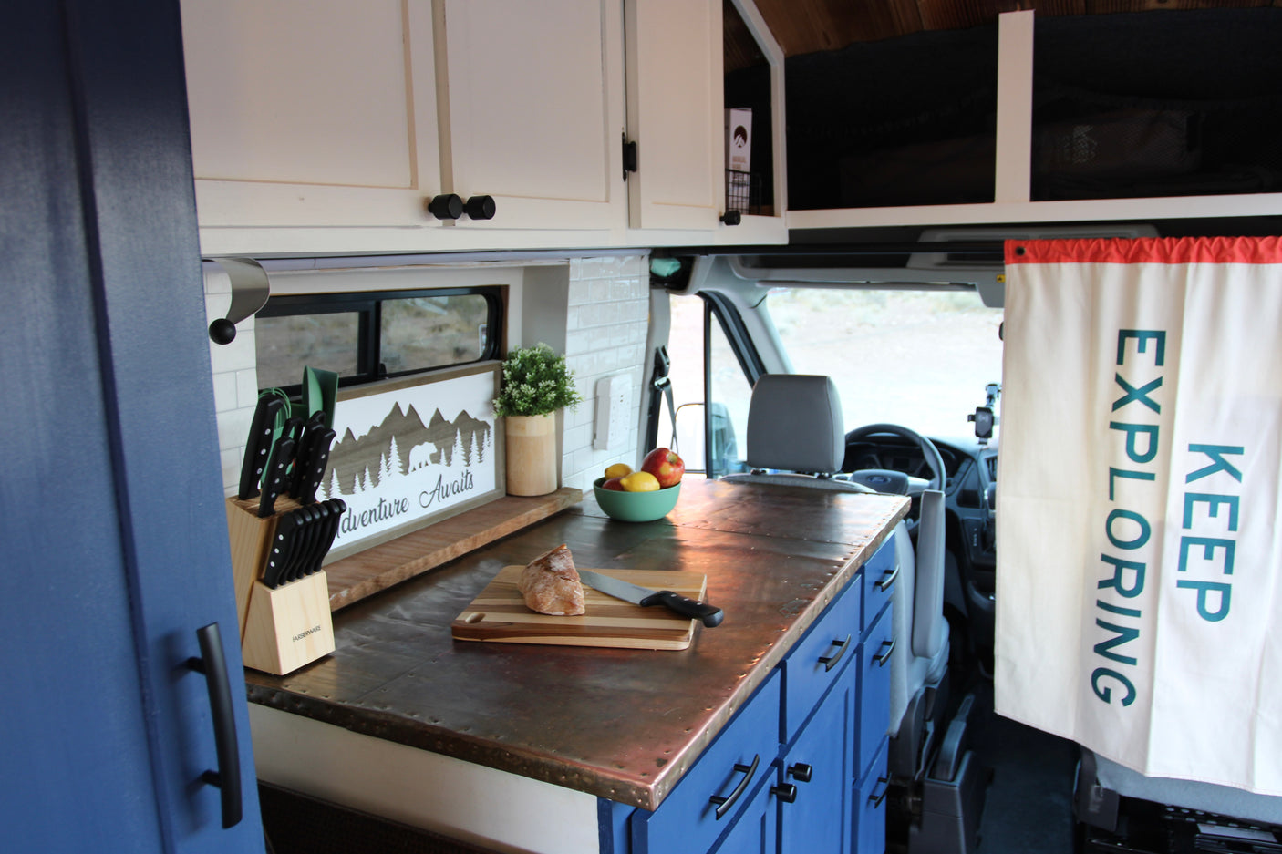 A compact RV rental kitchen boasts white and blue cabinets, a unique metal countertop, and 'Adventure Awaits' decor.