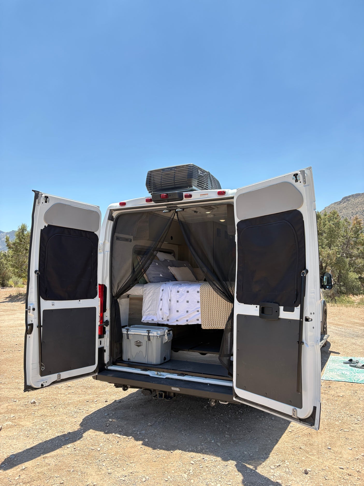 White RV rental's rear open, revealing a comfy bed, cooler, and mosquito net. Ideal for van life adventures.