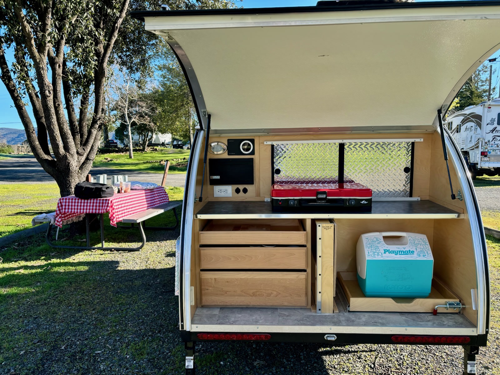An rv rental teardrop camper's open galley with stove, cooler, and drawers, ready for outdoor cooking by a picnic table.
