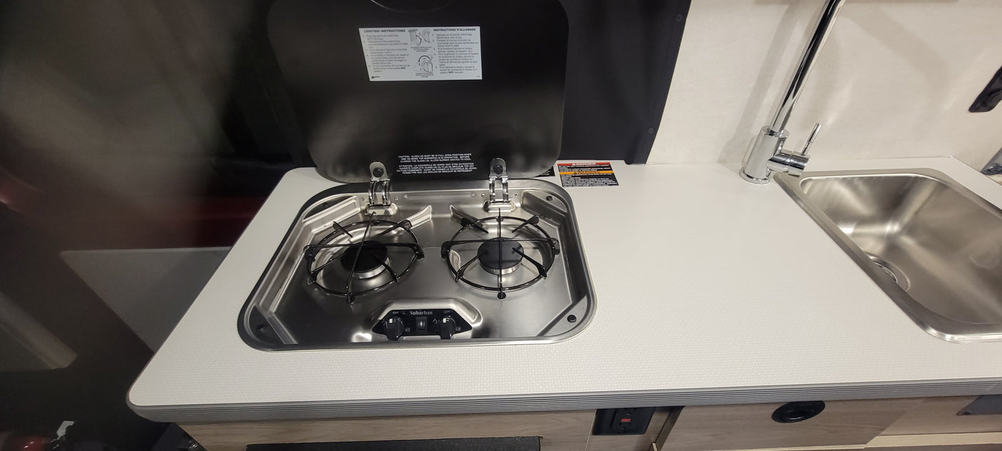 Inside an RV rental: a compact kitchen with a two-burner gas stove (Suburban) and stainless steel sink. Ideal for travel.