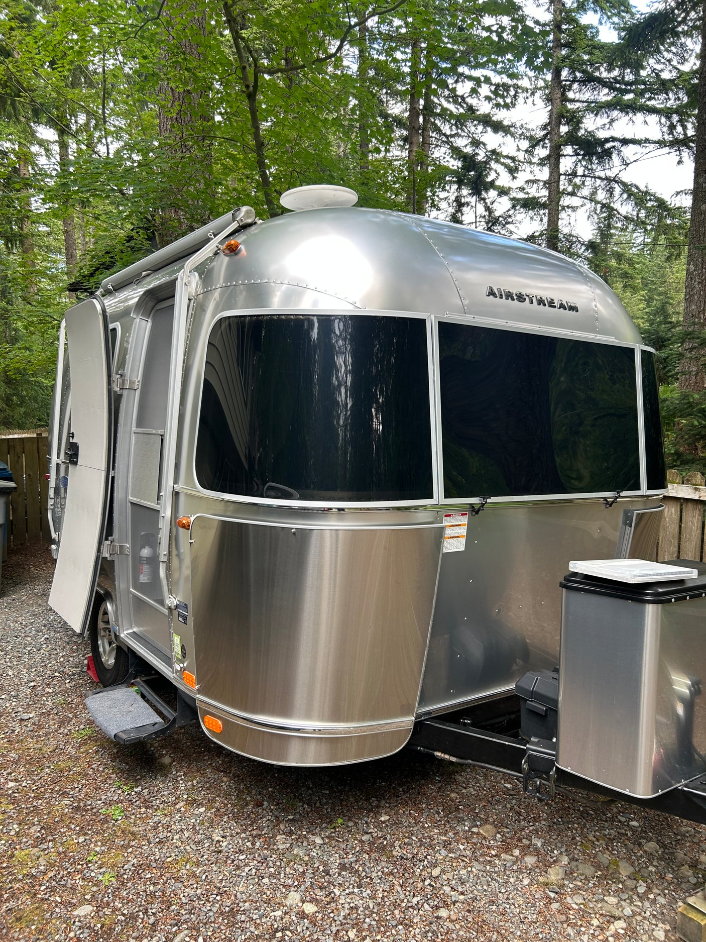 This gleaming Airstream travel trailer, ideal for an RV rental, is parked on gravel among tall trees, with its door open.