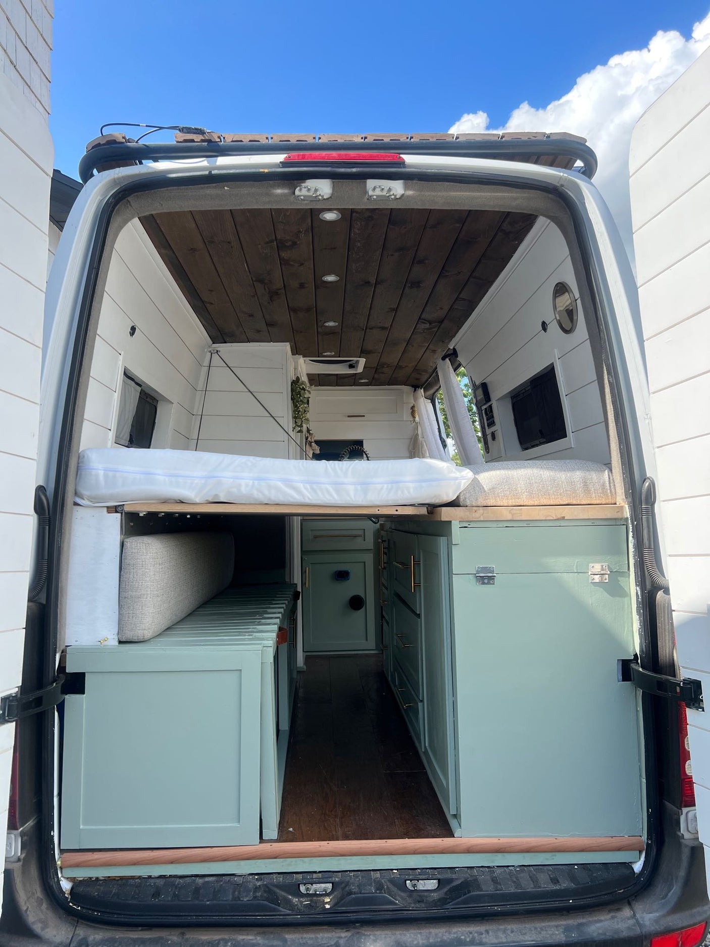 View into an RV rental van from its open back doors, showcasing the raised bed, wood ceiling, and light green cabinetry.