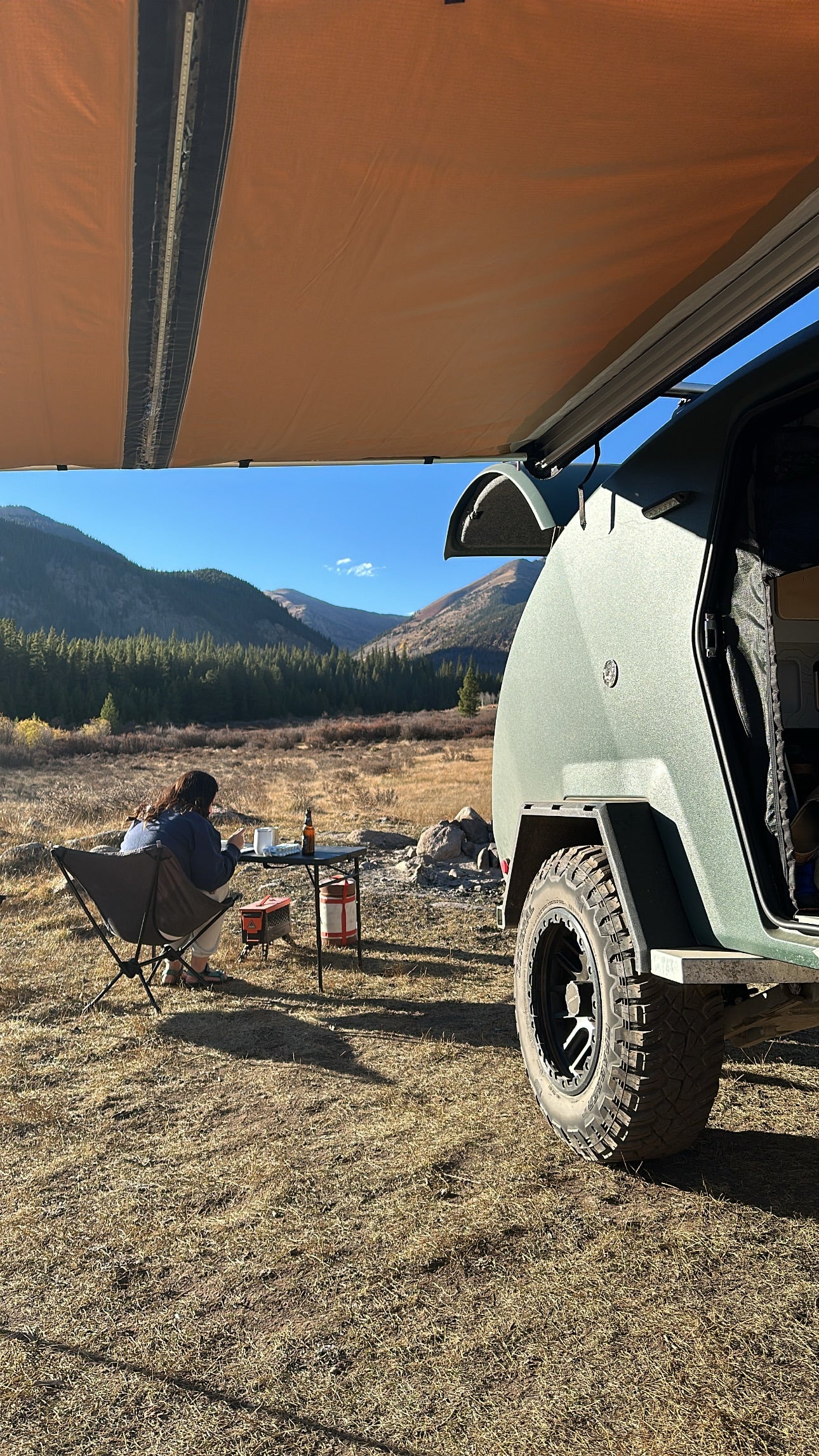 Camping with an RV rental: Person enjoys a meal beside a green teardrop trailer and awning in a mountain valley.