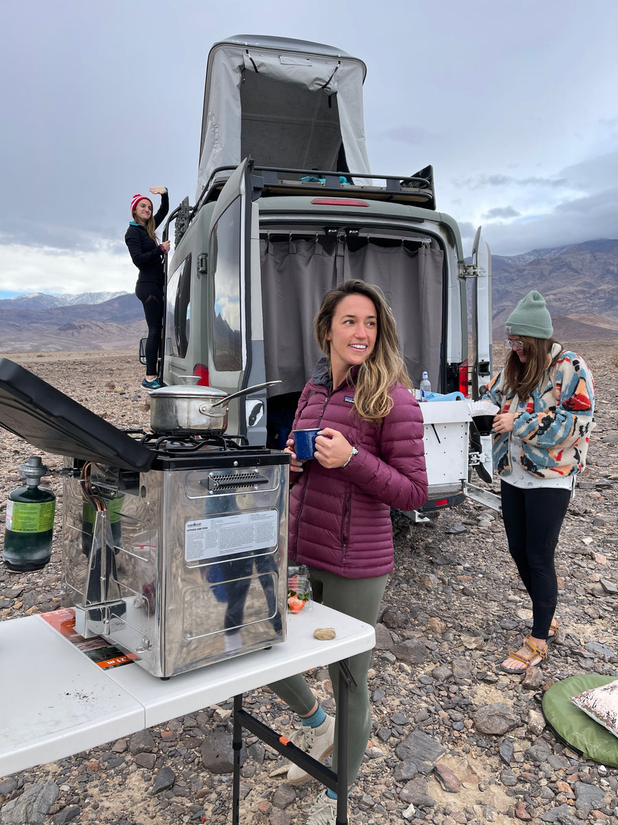 RV rental trip! Friends enjoy outdoor cooking & coffee by their camper van. One waves from the ladder, mountains behind.