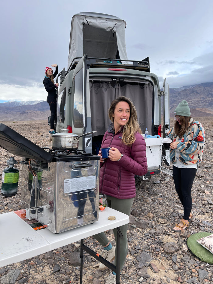 RV rental trip! Friends enjoy outdoor cooking & coffee by their camper van. One waves from the ladder, mountains behind.