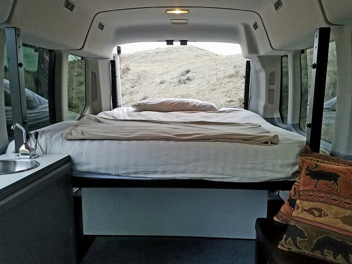 Cozy RV rental interior with a bed, small sink, and open back doors revealing a dry, grassy hill.