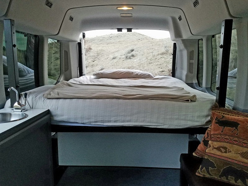Cozy RV rental interior with a bed, small sink, and open back doors revealing a dry, grassy hill.