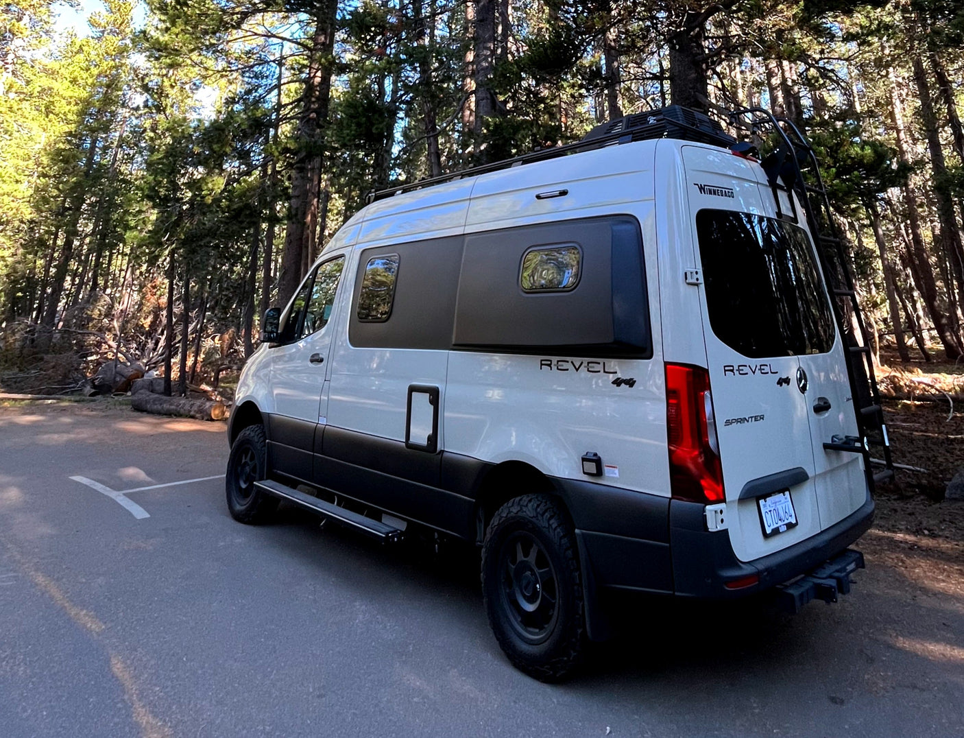 White Winnebago Revel 4x4 RV rental parked on asphalt, surrounded by tall pine trees, ready for adventure.