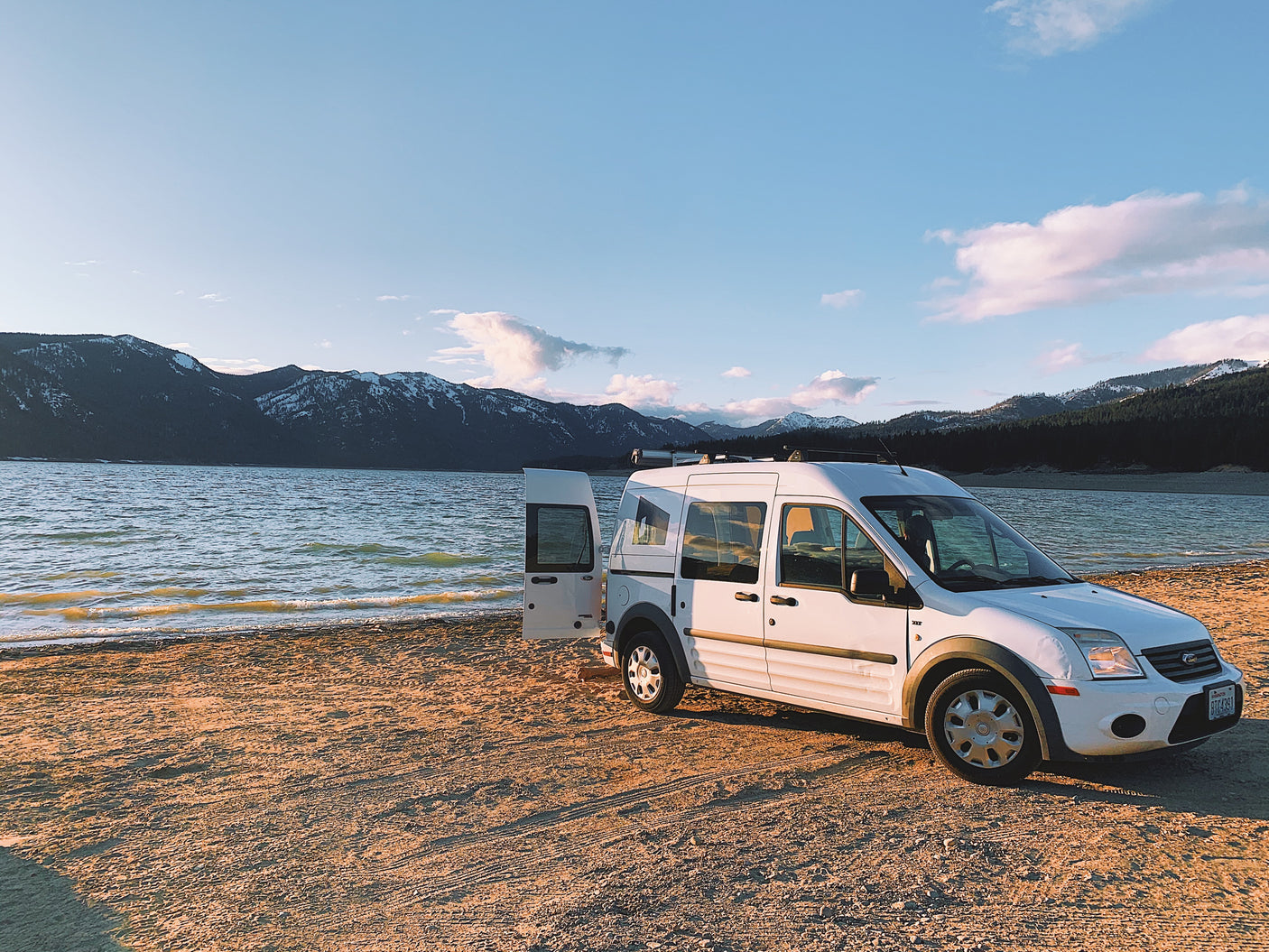 White RV rental van with open door on sandy shore, serene lake & snowy mountains in sunset light.