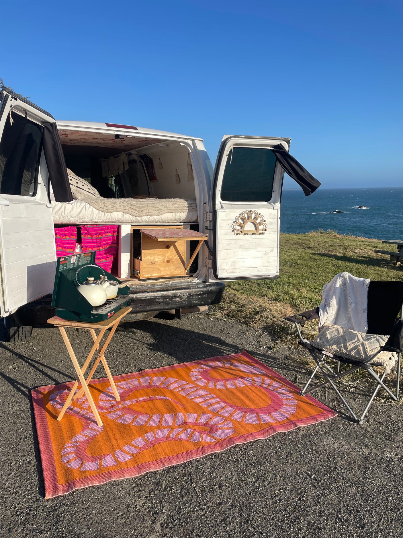 RV rental camper van parked by the ocean with an outdoor cooking setup, chair, and open doors showing the cozy interior.