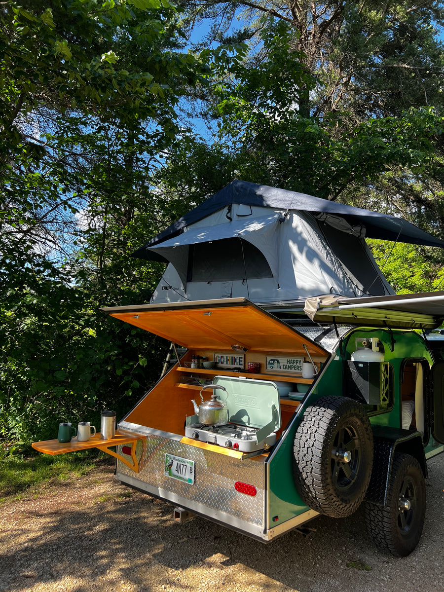 This RV rental teardrop trailer features a rooftop tent & outdoor kitchen, ready for camping in the woods.
