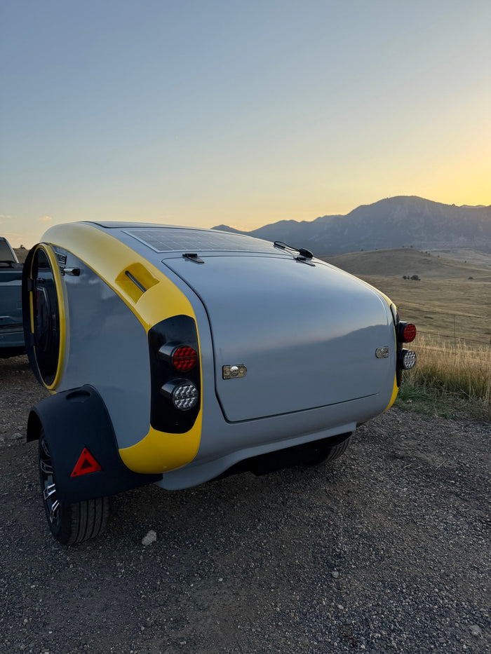 Modern gray & yellow travel trailer with solar panels, ideal for an RV rental adventure, parked on a gravel road with mountains at sunset.